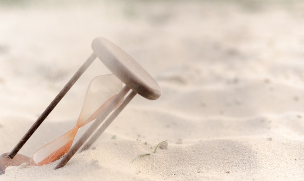 an hourglass rests in the sand, at a slight angle. there is a hazy tint to the air making it hard to see anything else.